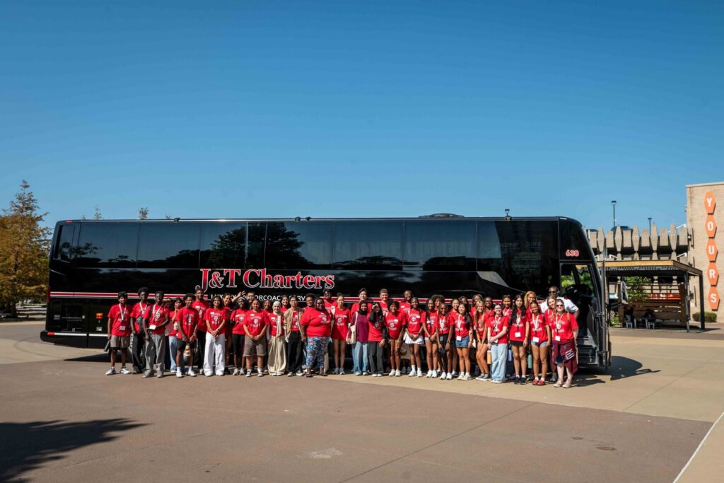A group of students from Rhodes College stand in front of a charter bus